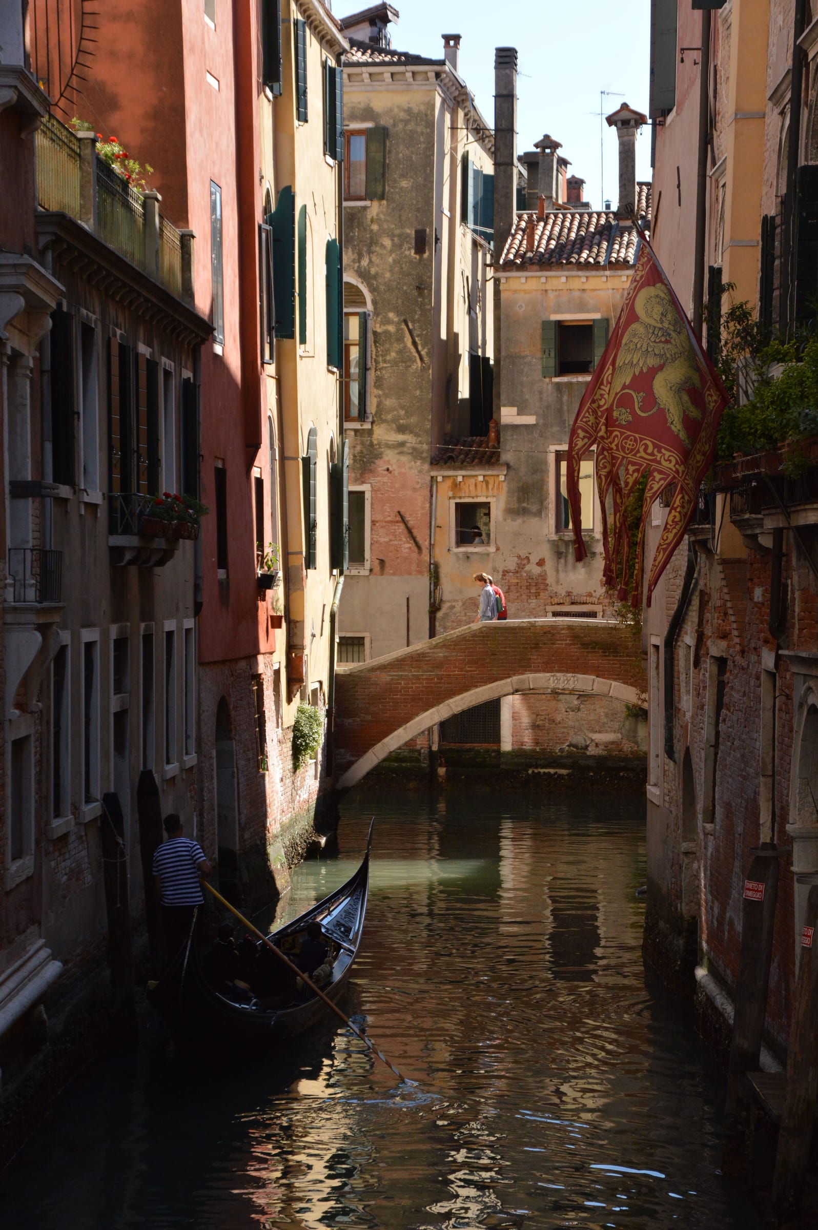 A Venice canal with a brick bridge and the red-and-gold flag of the Lion of Saint Mark